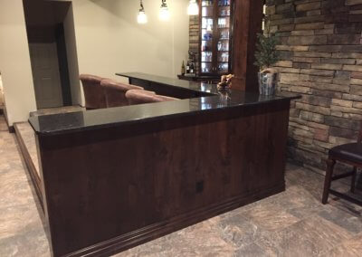 This was a project that we framed as well as the cabinetry. We used Alder wood with a toffee color stain on the cabinets, wainscoting, column bases, and wood beams. The cabinets are lit in the back bar. There is crown moulding on both the bar and serving area. The elevated area at the bar is very special to the owners. They wanted lounge chairs instead of stools.